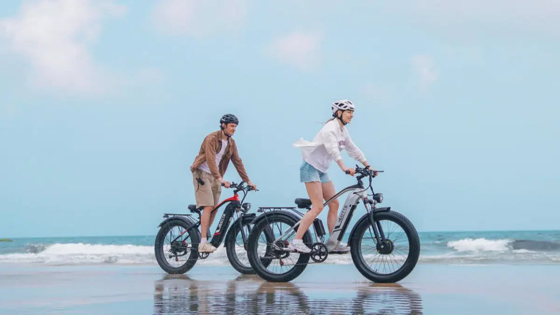  Two riders on e‑bikes cycling along a bright coastal beach under a clear blue sky, reflecting freedom, movement, and connection in natural daylight.