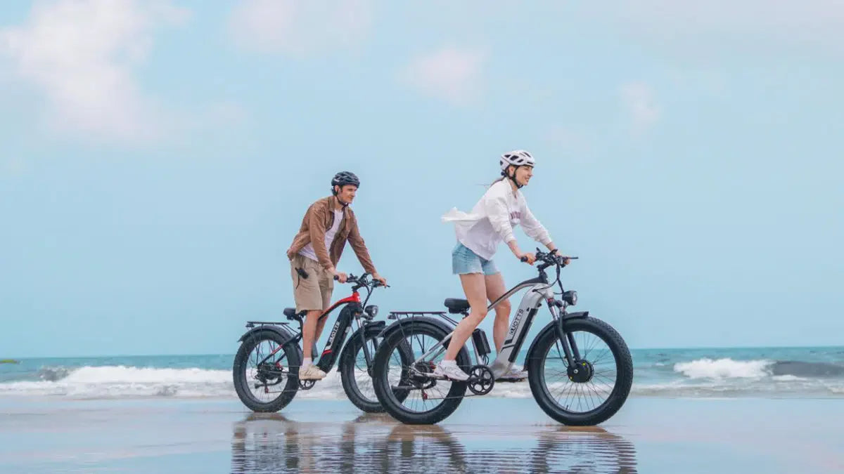  Two riders on e‑bikes cycling along a bright coastal beach under a clear blue sky, reflecting freedom, movement, and connection in natural daylight.