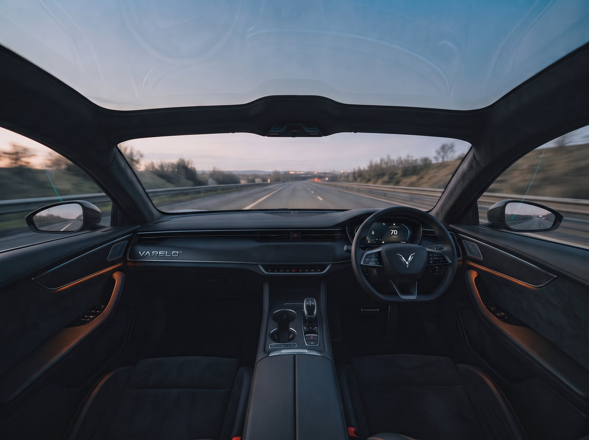 VARELO™ luxury electric vehicle obsidian cockpit with matte carbon-fibre trim, illuminated dashboard logo, and 70 mph digital cluster cruising a UK motorway at dusk.