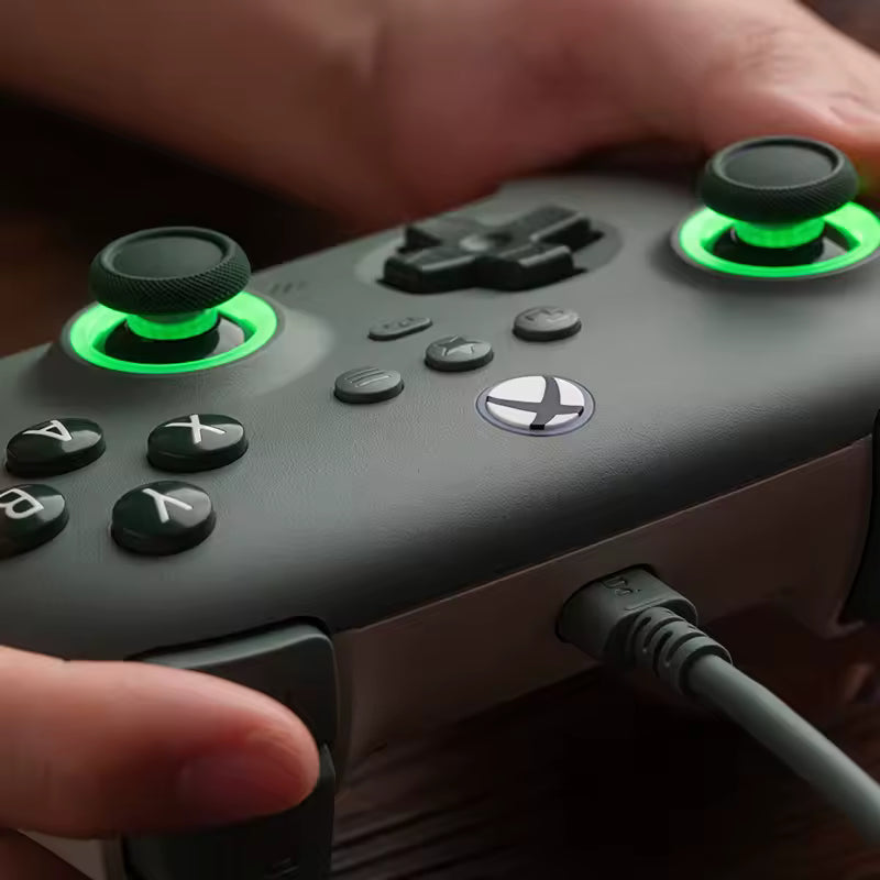 A close-up of a person holding a dark green Xbox wireless controller with glowing green thumbsticks, with a cable plugged into the side port.