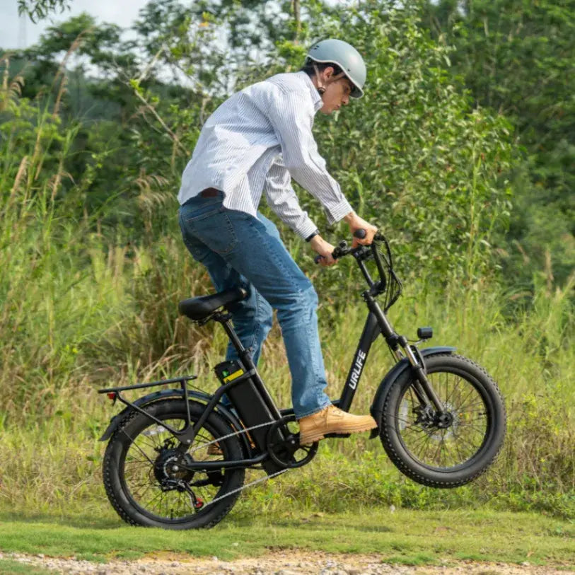 A man wearing a light blue helmet, white long-sleeve shirt, and blue jeans rides a black fat-tire electric bicycle outdoors on a grassy path with trees in the background.