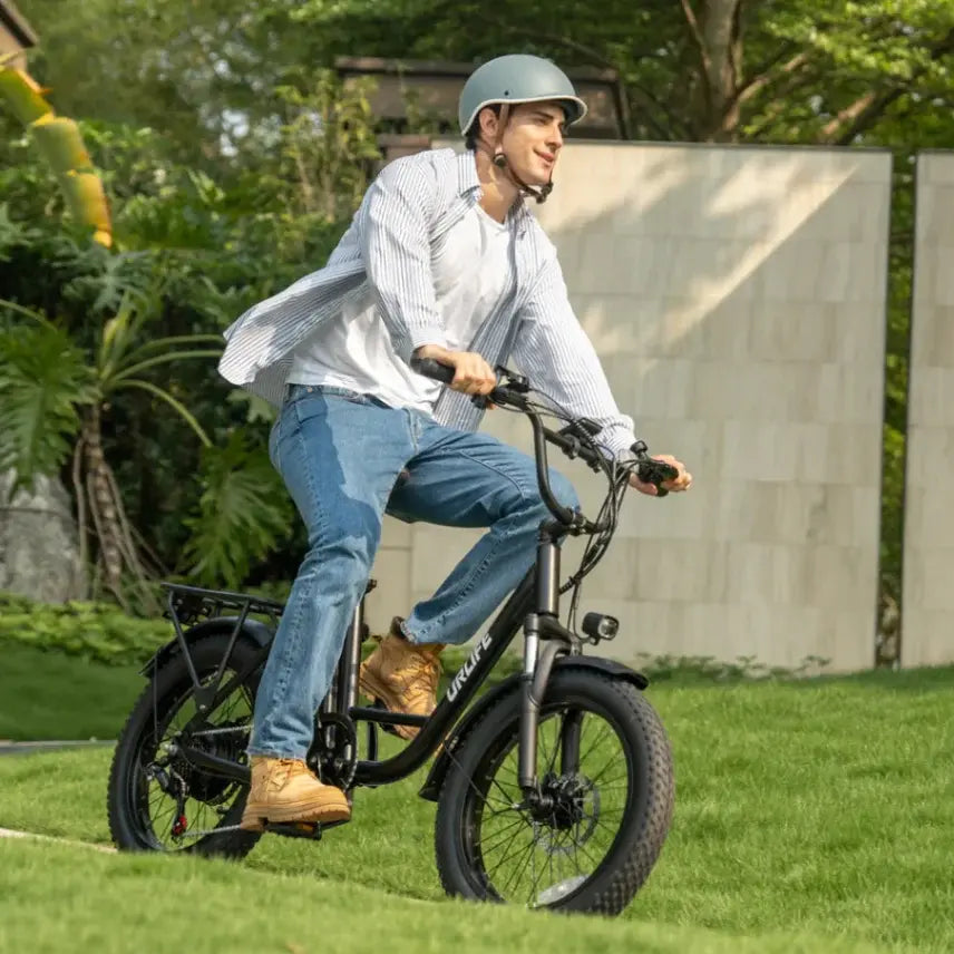 A man wearing a light blue helmet, a white striped button-down shirt, and blue jeans rides a black electric bicycle on a green grassy lawn with a stone wall in the background.