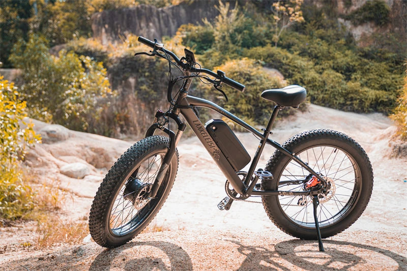Black LANKELEISI MG740PLUS full suspension fat tyre electric mountain bike parked on a rocky trail surrounded by trees and vegetation under natural daylight, showing 26x4.0 inch tyres and central battery frame.
