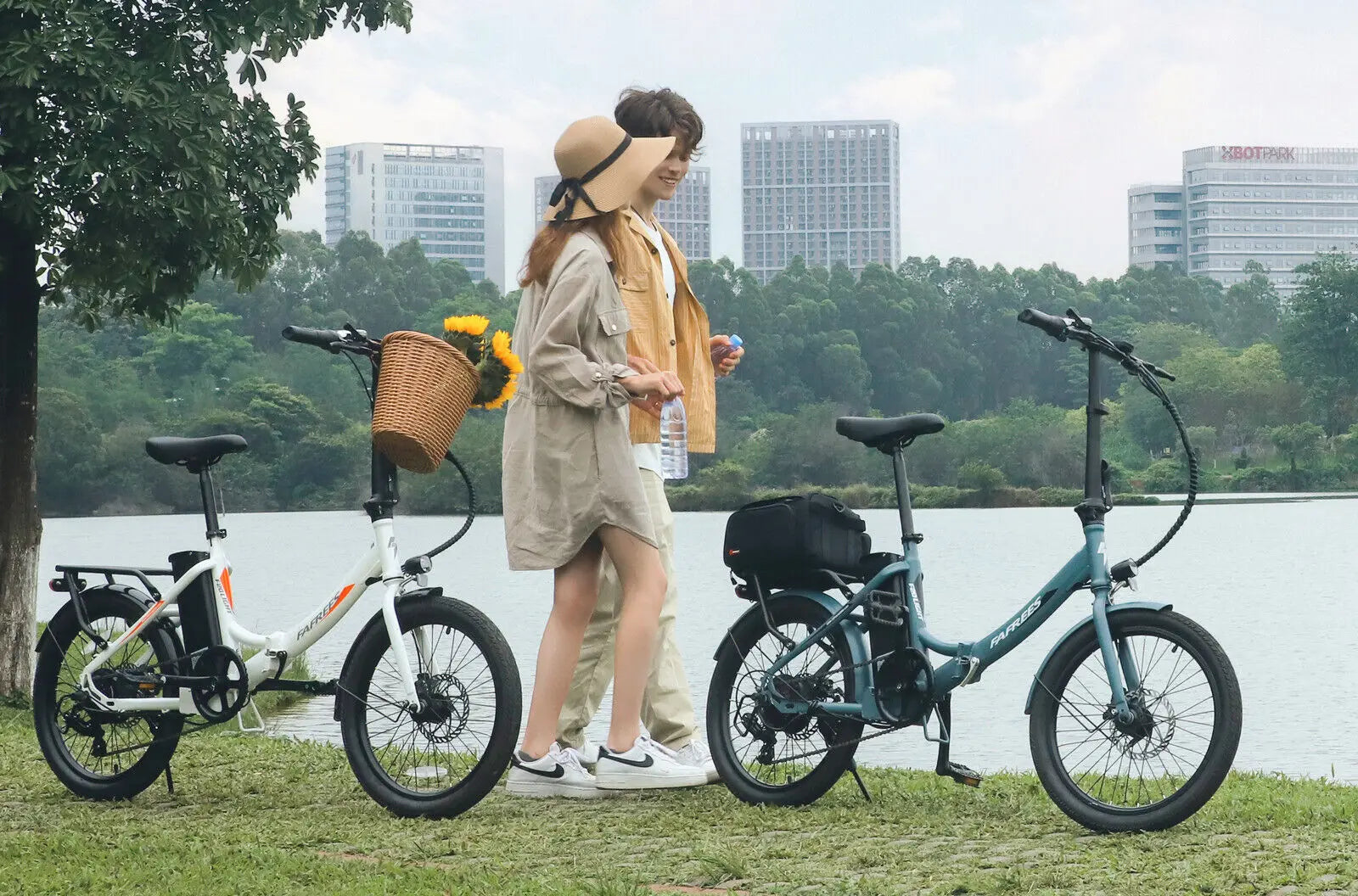 Two electric bicycles parked by a body of water with two people standing between them, surrounded by greenery and buildings.