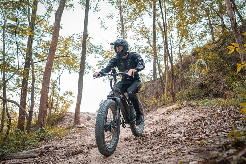 Rider wearing protective gear riding a LANKELEISI MG740PLUS full suspension fat tyre electric mountain bike through a forest trail with autumn leaves and trees.