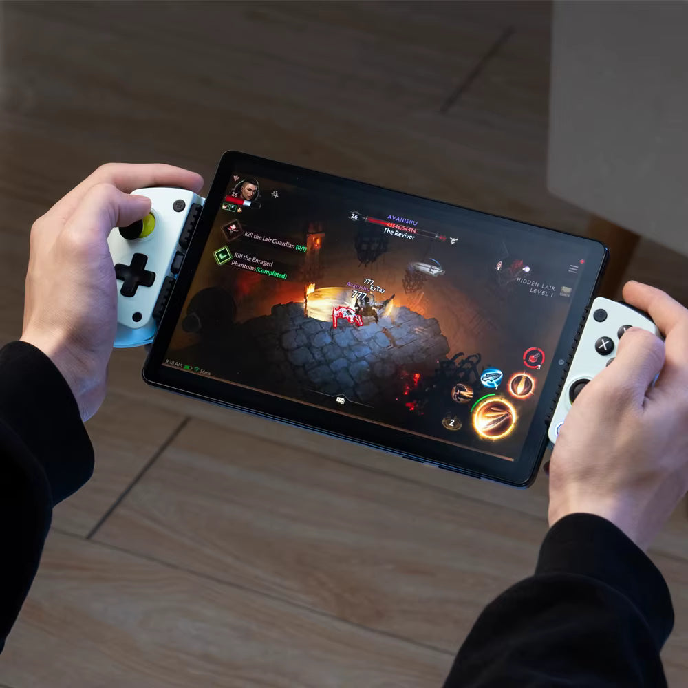 A person holding a tablet game controller setup with two wasaibi coloured game controllers and a black tablet showing a video game on screen, photographed from above on a wooden floor.
