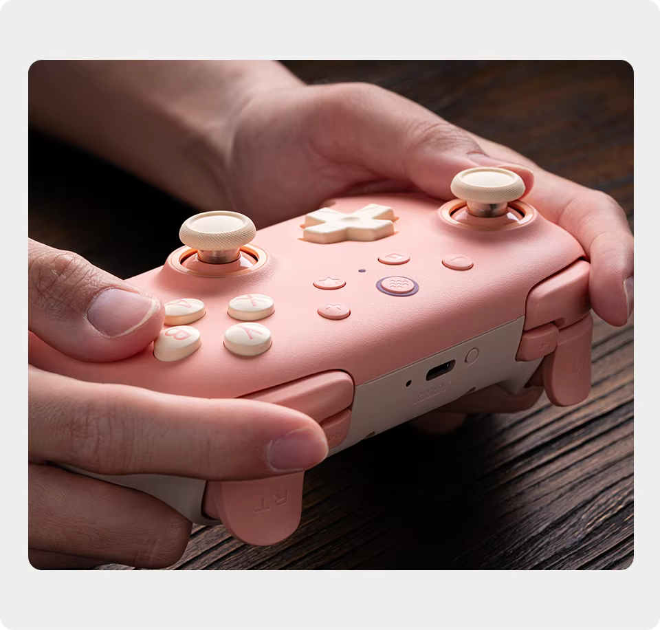 Close-up of hands holding the 8BitDo Ultimate 2C Wireless Controller in Sunset Peach showing ergonomic grip, Hall Effect joysticks, and custom shoulder bumpers