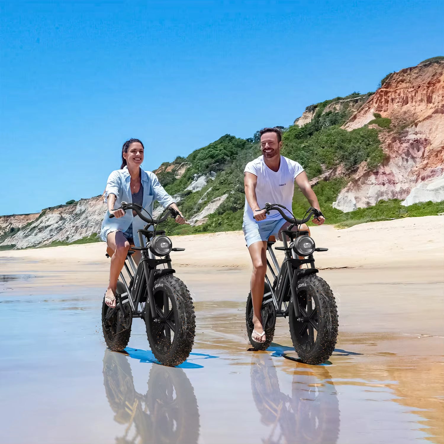 Two people riding JANSNO X50 e-bikes on a beach. The electric bikes are JANSNO X50 on a sandy beach with cliffs in the background.