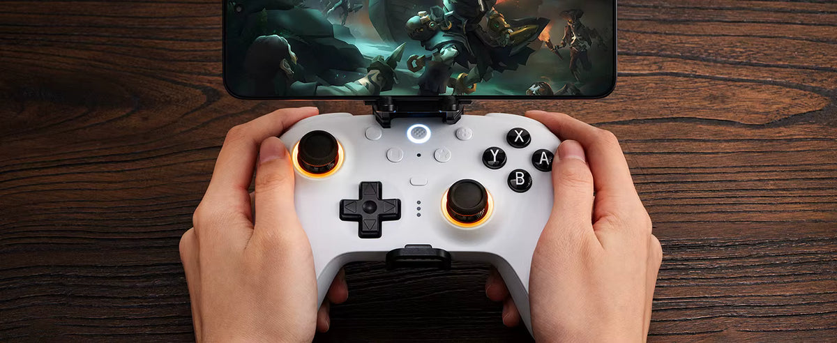 8BitDo lifestyle product photo showing a white wireless controller paired with a mobile phone mount clip, displaying an active gaming session on the smartphone screen over a rustic wooden desktop background.