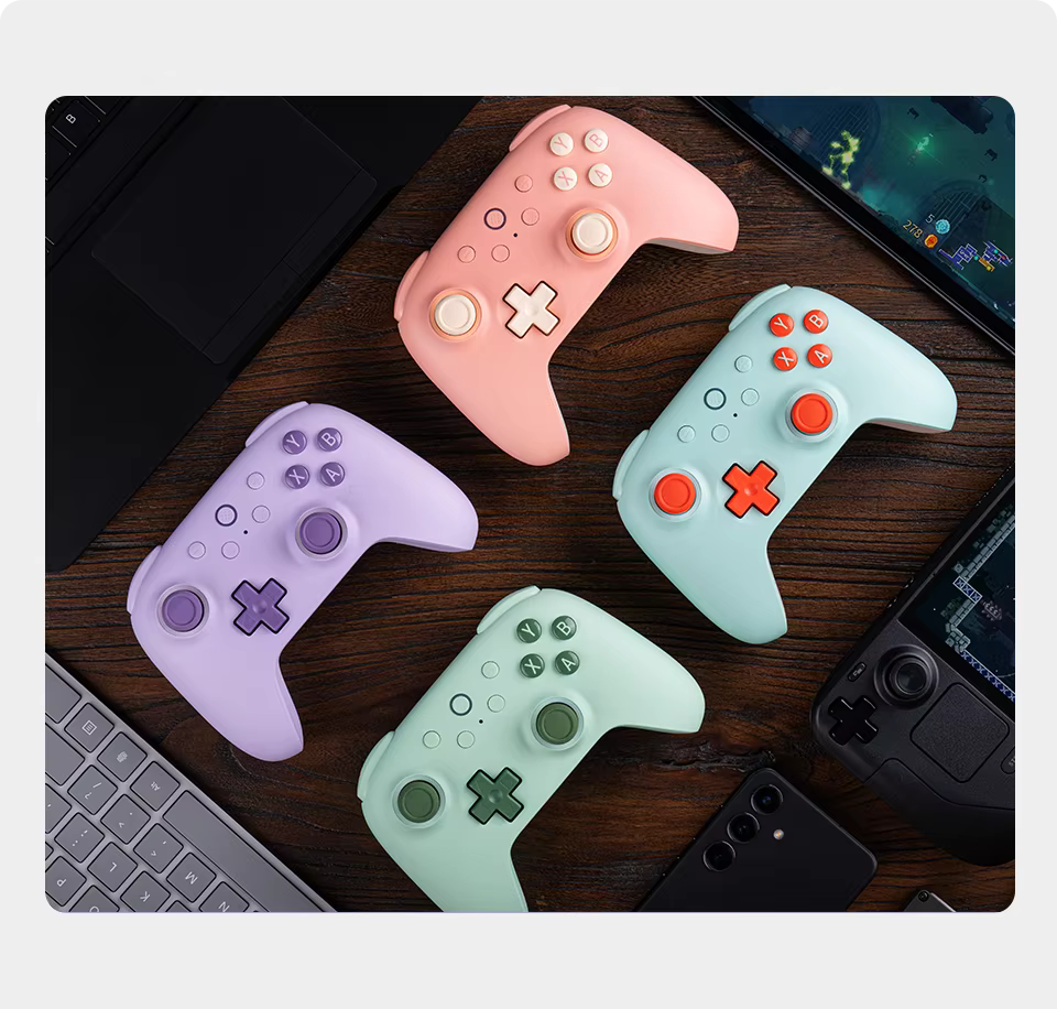 Four colorful gaming controllers on a wooden surface with a laptop and phone in the background.