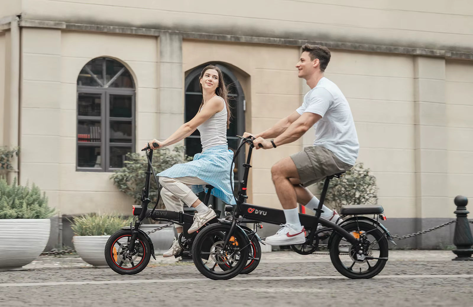 Two people riding DYU A1F Pro Folding E-Bike  electric bikes on a street with a building in the background