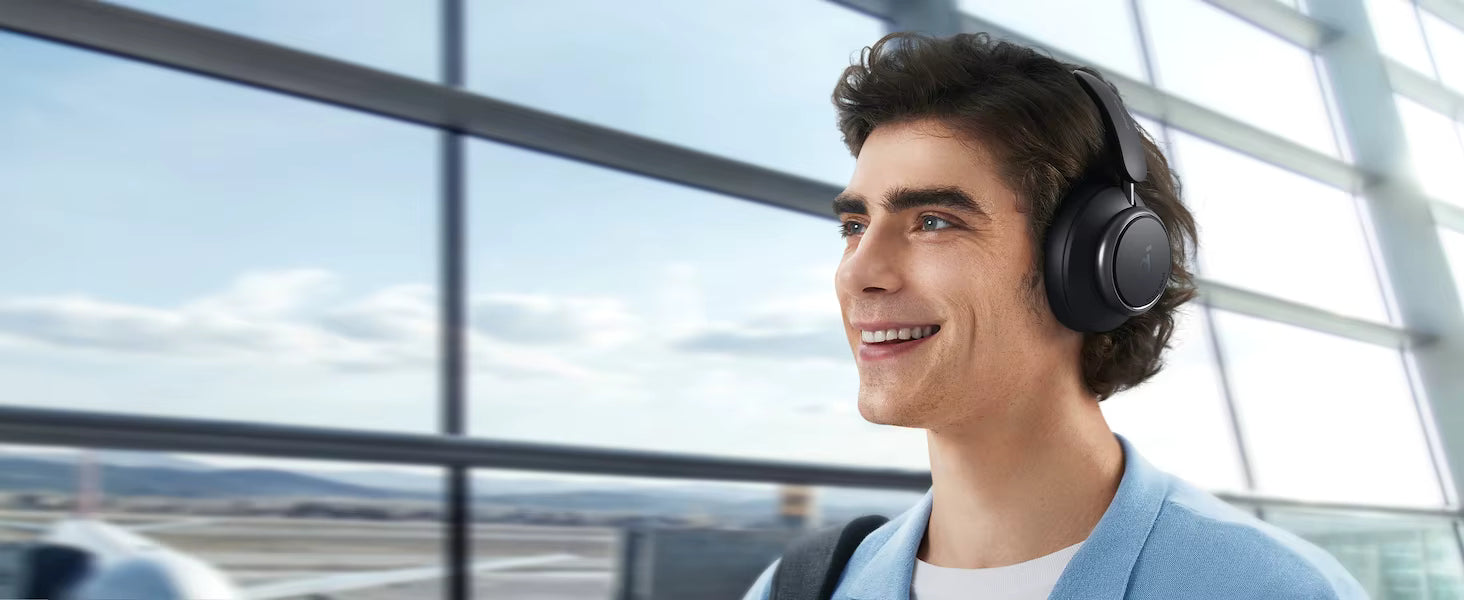A man wearing black over-ear headphones stands in an airport terminal with large windows, looking out toward the tarmac under a cloudy sky.