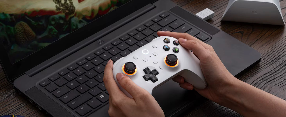 Lifestyle action shot of a white 8BitDo Ultimate 2 Wireless Controller being held by a gamer in front of an open gaming laptop on a wooden desk, featuring glowing orange RGB fire rings around the joysticks.