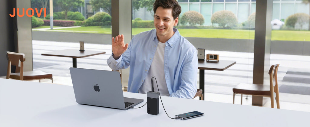 A man in a cafe setting waves while using a silver laptop connected to a black JUOVI power bank, with a smartphone nearby on a white table.
