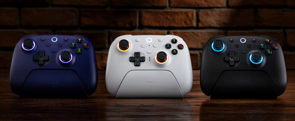 Studio showcase of the complete 8BitDo Ultimate 2 Wireless Controller lineup resting on their respective charging docks against a dark brick wall background, featuring three colorways: dark blue on the left, classic white in the center, and matte black on the right.