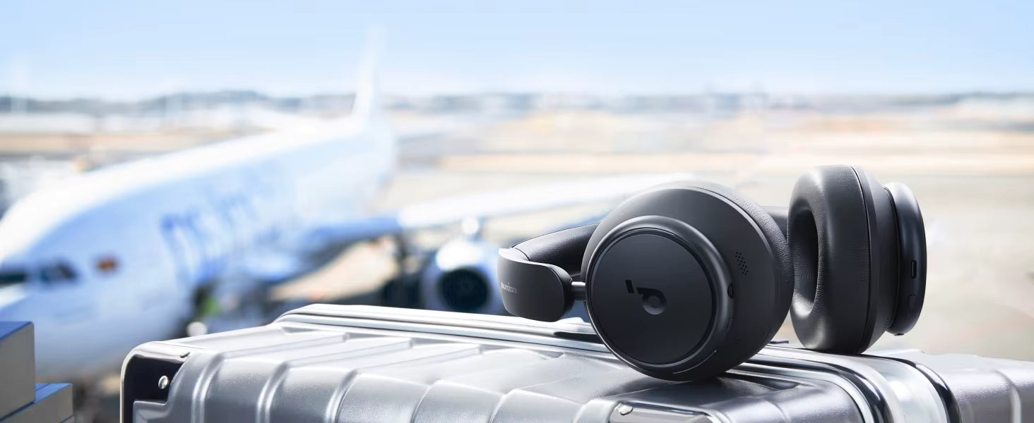 Black over-ear headphones resting on a gray hard-shell suitcase, with a blurred airplane and airport tarmac in the background.