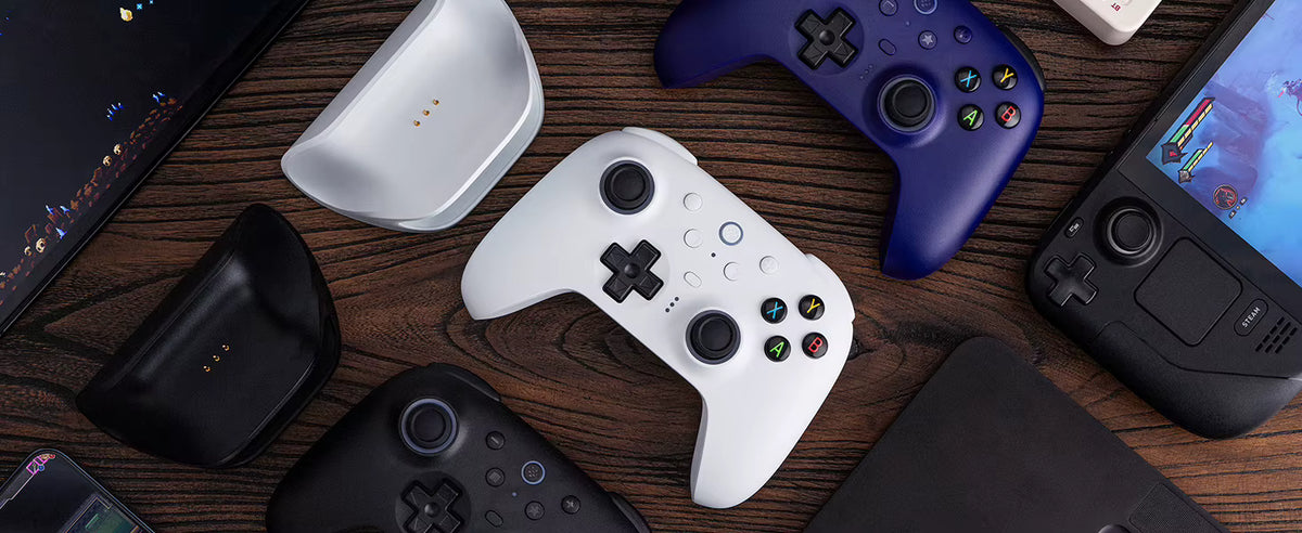 Flat lay lifestyle product shot featuring the VARELO™ e-commerce tech setup, including a white 8BitDo Ultimate 2 Wireless Controller centrally placed, magnetic charging docks in white and black, a blue controller variant, a handheld gaming console, and accessories arranged on a rustic wooden desktop.