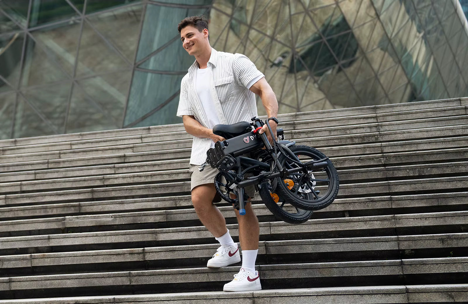 Man holding a folded DYU A1F Pro Folding Electric Bike e-bike on outdoor steps with modern architecture in the background