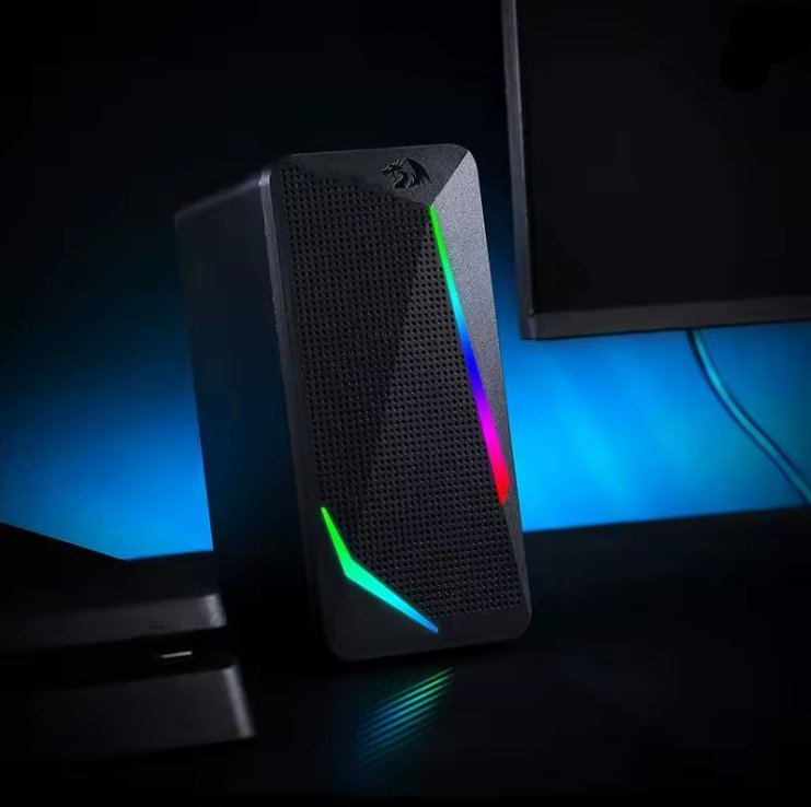 A black gaming desktop speaker with RGB LED light accents on the front, shown in a dark room setting with blue ambient lighting.