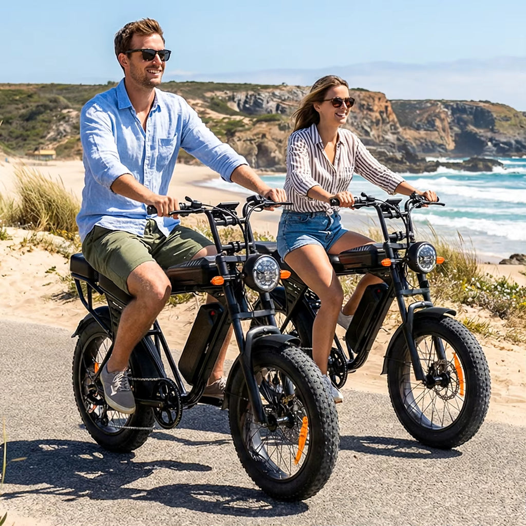 Two people riding electric bikes on a coastal road with ocean and cliffs in the background.