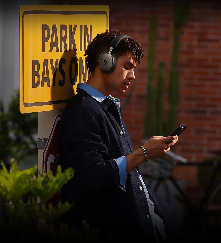 Person wearing Baseus Inspire XH1 headphones and looking at a phone next to a 'Park in Bays On' sign.