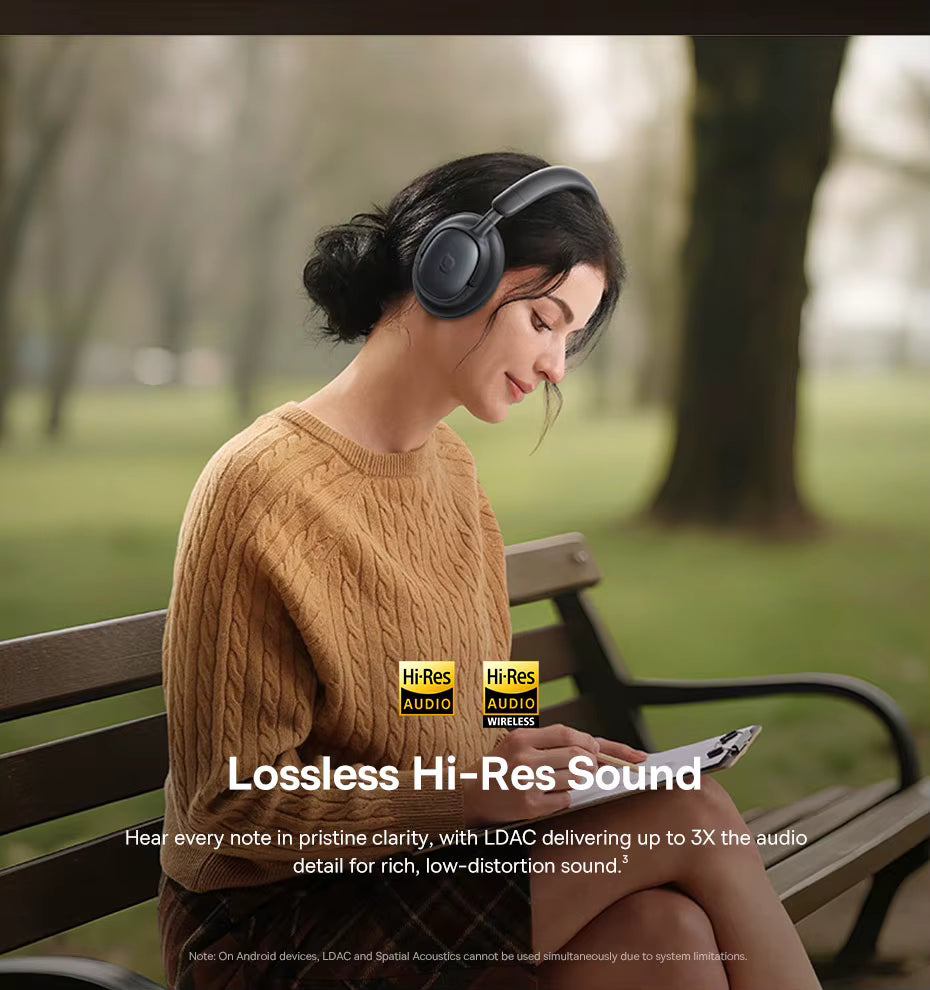 A woman wearing dark gray over-ear headphones sits on a park bench, looking down at a phone. The background is a softly blurred green park with trees.