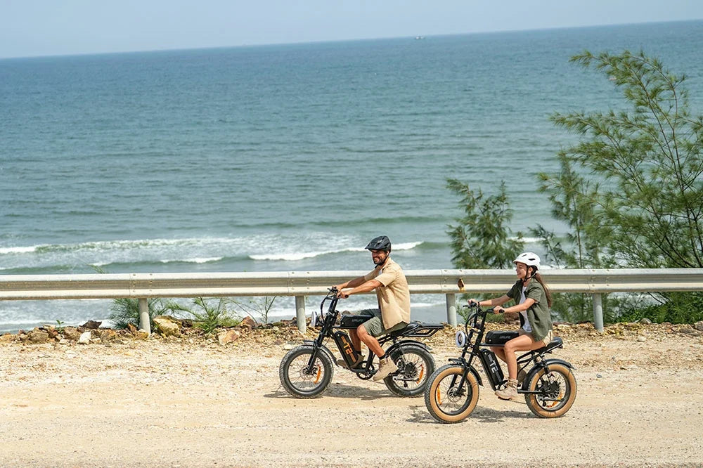 Two people riding URLIFE T2 electric bicycles on a sandy coastal road with the ocean and guardrail in the background.