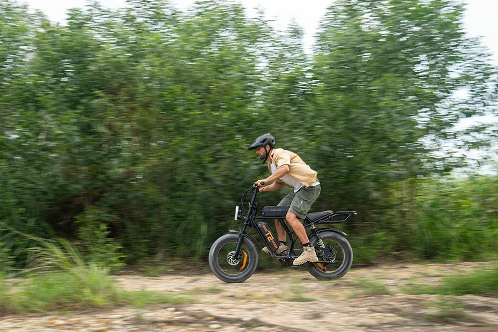 A man riding a URLIFE T2 electric fat-tire bicycle on a dirt path with a blurred green forest background.