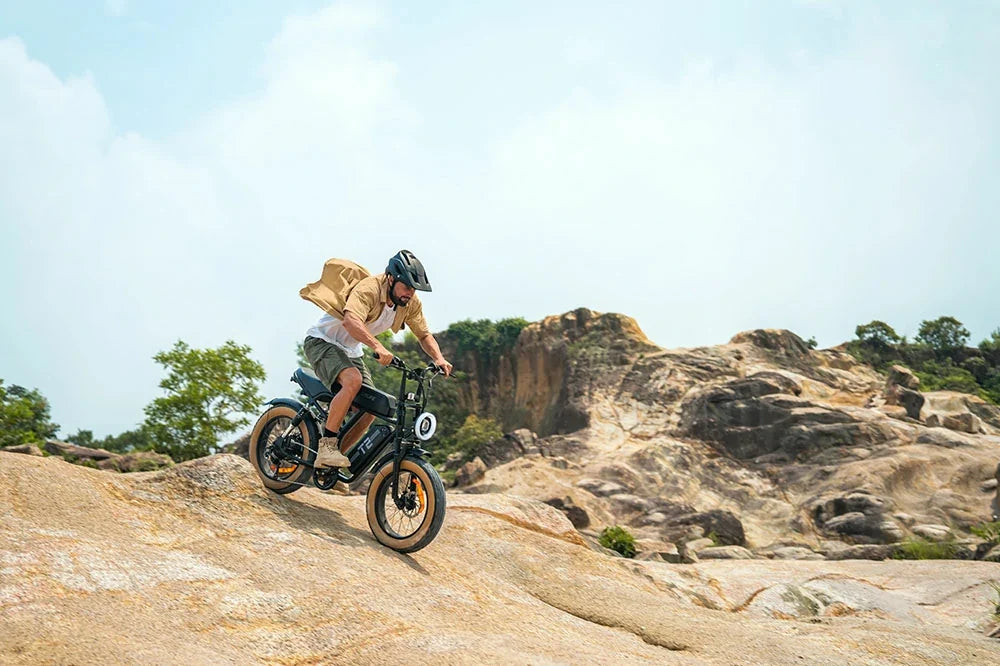 A person riding a black URLIFE T2 electric e-bike with gold accents up a rocky sandy path in an outdoor desert landscape under a cloudy sky.