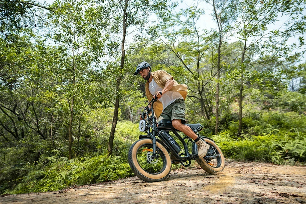 A man riding a T2 electric fat-tire bike on a dirt path surrounded by a dense green forest. The rider is wearing a helmet, a tan jacket, and green shorts.